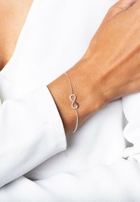 Pulsera de plata con un símbolo de infinito, unida por una delicada cadena, exhibida en una muñeca bronceada sobre un fondo blanco.