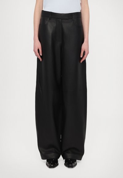 Schwarze Leder-Hose mit weitem Bein und glatter Textur, mit einem Gürtel in der Taille und Taschen. Getragen mit schwarzen Schuhen.