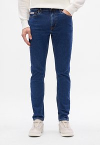 Ciemnoniebieskie dżinsy slim fit z bawełny, z subtelnym przetarciem, pięć kieszeni i kontrastowe szycie. W zestawieniu z jasnymi trampkami.