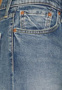 Pantalones vaqueros de denim azul claro con remaches de cobre, costuras naranjas, un bolsillo delantero y un ligero desgastado, enfatizando un diseño casual.