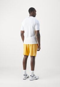 Weißes Sportshirt mit kurzen Ärmeln und gelben Shorts. Das Shirt hat eine taillierte Passform und ein Logo auf der Rückseite, während die Shorts leicht sind.