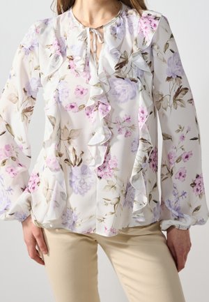 Kvinde iført en hvid bluse med lange ærmer med pink og lilla blomstermønster og flæser foran kombineret med beige bukser.