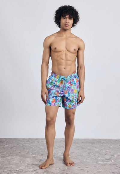 Tânăr cu păr creț, îmbrăcat în șorturi de baie colorate și tipărite, stând desculț pe un podea gri, în fața unui zid simplu de culoare deschisă.