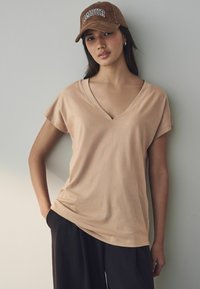 Beżowa koszulka typu v-neck z krótkim rękawem, o miękkiej teksturze, w zestawie z luźnymi ciemnymi spodniami. Dodatki to brązowa czapka z kordurowego materiału z napisem "AMOUR".