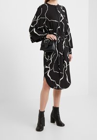 Schwarzes Kleid mit weißem abstraktem Muster, lange Ärmel, tailliert, seitliche Schlitze. Kombiniert mit einer schwarzen Umhängetasche und schwarzen Stiefeletten.