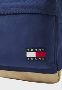 Tamsiai mėlynas audinio kuprinė, žemiau kurios yra grietinėlės spalvos dalis, papuošta juodu stačiakampiu ženkliuku, ant kurio užrašyta "TOMMY JEANS" su spalvotais akcentais.