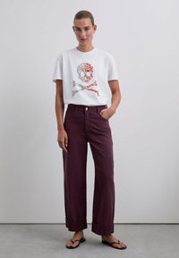 Camiseta blanca con un gráfico de calavera y tibias cruzadas, combinada con pantalones anchos burdeos y chanclas, con puños enrollados.