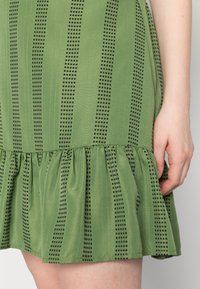 Vestido verde com padrões xadrez pretos, um tecido texturizado e uma bainha com volantes. Detalhe da saia e da colocação da mão.