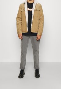 Beżowa kurtka z korduroju z białym kołnierzem z owczej wełny, czarny T-shirt z grafiką, szare spodnie oraz czarne buty na sznurówki. Prosty design, bez wzorów.