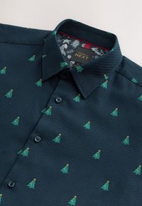 Chemise boutonnée vert foncé avec de petits sapins de Noël brodés et un col intérieur à motifs.
