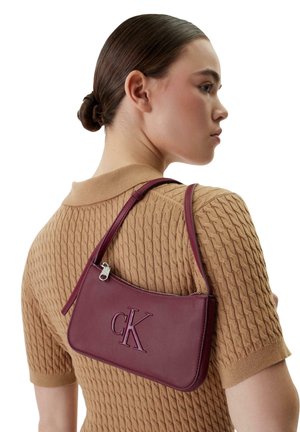 Femme aux cheveux tirés en arrière portant un haut texturé à manches courtes de couleur beige, portant un sac à bandoulière Calvin Klein bordeaux avec une fermeture éclair visible.