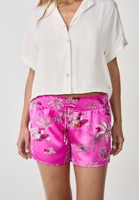 Roze satijnen shorts met een smockband, versierd met een bloemenprint in wit en paars. Gecombineerd met een witte button-up shirt.