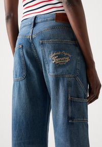 Blue denim jeans featuring a straight-leg design, two back pockets, and embroidered "Tommy Jeans" logo in gold on the right pocket.