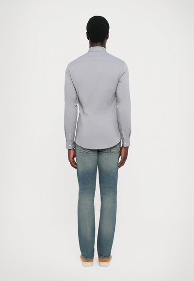 Lys grå langermet skjorte med knappede mansjetter, kombinert med falmede blå rette jeans og lyse beige sko, sett fra baksiden.
