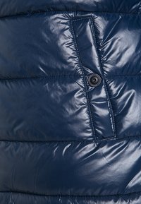 Námořnická modrá péřová bunda se lesklou, prošívanou texturou. Obsahuje prošívanou kapsu a černý ozdobný patent.
