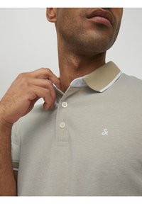 Polo shirt gris clair avec un col beige à accents blancs, deux boutons et un petit logo blanc sur la poitrine, tissu texturé.