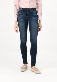Dark blue, fitted jeans made of denim. Features a high waist and subtle whiskering. Paired with beige ballet flats.