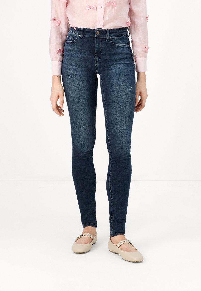 Dark blue, fitted jeans made of denim. Features a high waist and subtle whiskering. Paired with beige ballet flats.