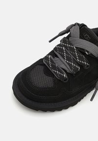 Baskets noires avec une tige en suede et des accents en maille, des lacets texturés gris et noirs, et une semelle en caoutchouc épaisse pour l'adhérence.