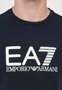 Marineblå bomull T-skjorte med et stort brodert hvitt logo med "EA7" og "EMPORIO ARMANI" tekst, rund hals og korte ermer.