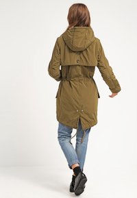 Mujer alejándose vestida con una parka con capucha de color verde oliva, jeans claros remangados, calcetines negros y zapatos negros.