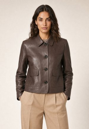 Femme aux cheveux ondulés foncés portant une veste en cuir marron foncé boutonnée et un pantalon beige à jambes larges, debout les mains dans les poches.