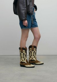 Braune und beige Cowboy-Stiefel mit filigranem, ausgeschnittenem Blumenmuster, aus Wildleder und Leder, spitze Zehen, niedriger Absatz und kontrastierende Akzente.