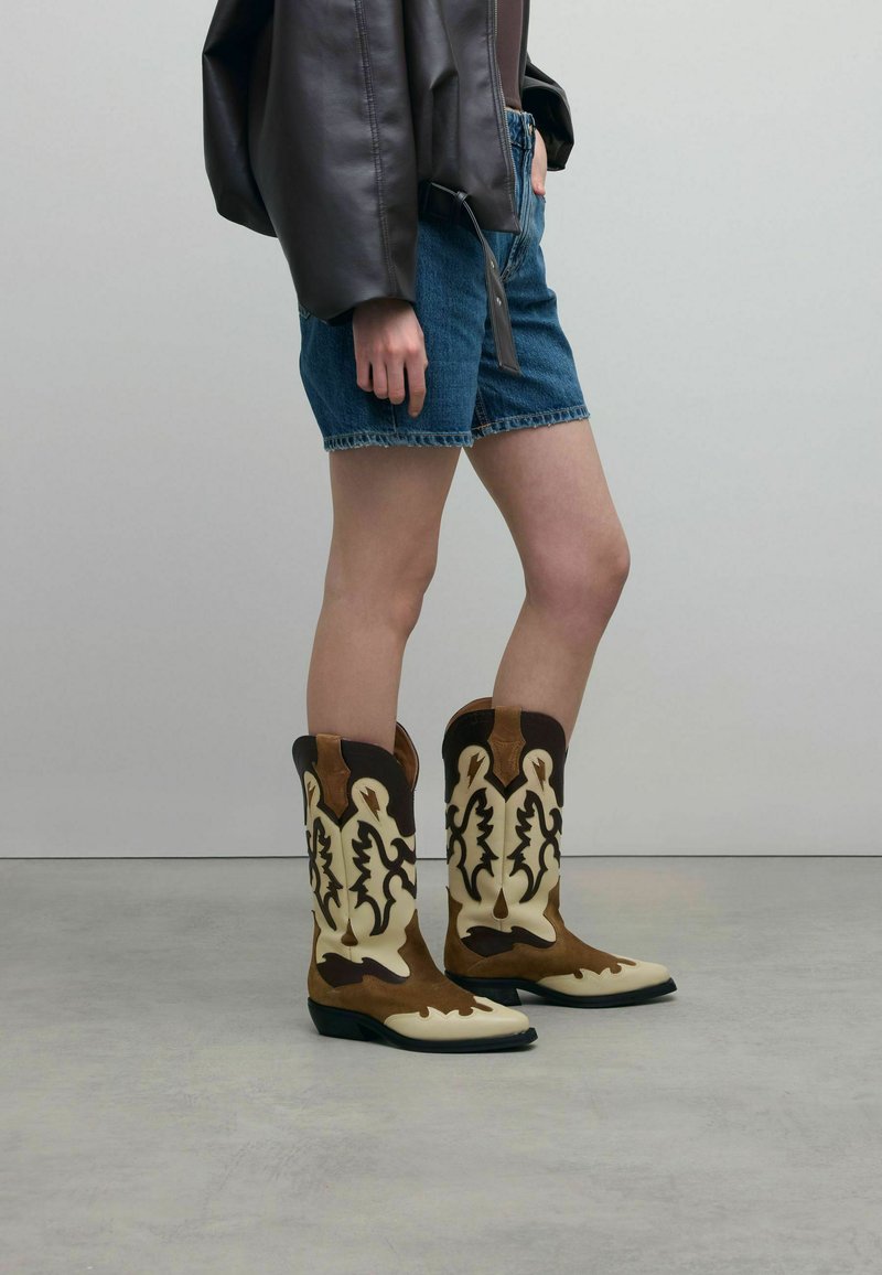 Braune und beige Cowboy-Stiefel mit filigranem, ausgeschnittenem Blumenmuster, aus Wildleder und Leder, spitze Zehen, niedriger Absatz und kontrastierende Akzente.