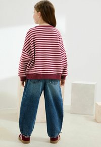 Sweatshirt maroon à rayures avec coupe ample, associé à un jean bleu ample. Détails comprennent des poignets et une taille côtelés, ainsi que des chaussures rouges.
