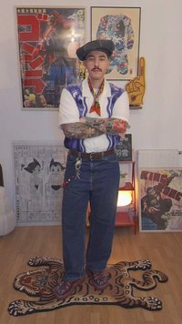 Homem com tatuagens e bigode, vestindo uma boina, uma camisa estampada e jeans, está de pé sobre uma pele de tigre numa sala com posters emoldurados.
