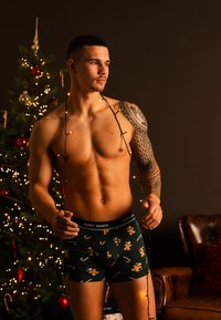 Boxershorts i mörkgrön med mönster av pepparkaksgubbe och godiskäppar; modellen har en tatuering och håller ljusslingor mot en festlig bakgrund.