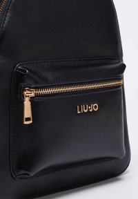 Sac à dos en cuir noir avec une poche avant zippée, ferrures dorées et logo de la marque en relief. Texture lisse avec un design structuré.