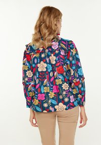 Blusa a maniche lunghe con motivo floreale blu e dettagli arricciati. Presenta vari fiori colorati, foglie e una vestibilità comoda. Abbinata a pantaloni beige.