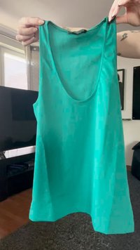 Delle mani tengono in alto una canotta senza maniche di colore turchese con scollo rotondo all'interno di un soggiorno con TV e pavimento coperto da tappeto.