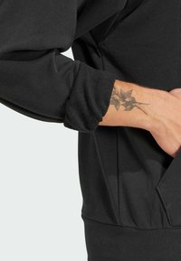 Crni pulover s dugim rukavima i teksturiranim manžetama, koji se ističe opuštenim krojem i bočnim džepovima. Vidljivi tetovaža lista na zapešću.