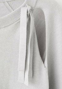 Pulover tricotat gri deschis cu design cu umerii decupați. Prezintă șnururi lungi din material și guler nervurat, evidențiind textura moale.