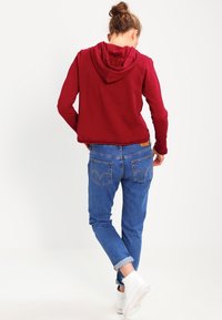 Sudadera con capucha color burdeos, de corte holgado, combinada con unos vaqueros de mezclilla azul. Los vaqueros tienen un dobladillo enrollado y una costura distintiva en los bolsillos traseros. Zapatillas blancas.