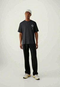 Mann mit schwarzem T-Shirt mit Sonne- und Mondgrafik, schwarzen Hosen, weißen Sneakern und hellgrauerm Cap, der vor einem schlichten Hintergrund steht.