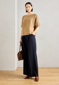 Top bej tricotat, cu mâneci scurte, asortat cu pantaloni largi cu dungi bleumarin. Gentă din piele maro și pantofi asortați completează ținuta.