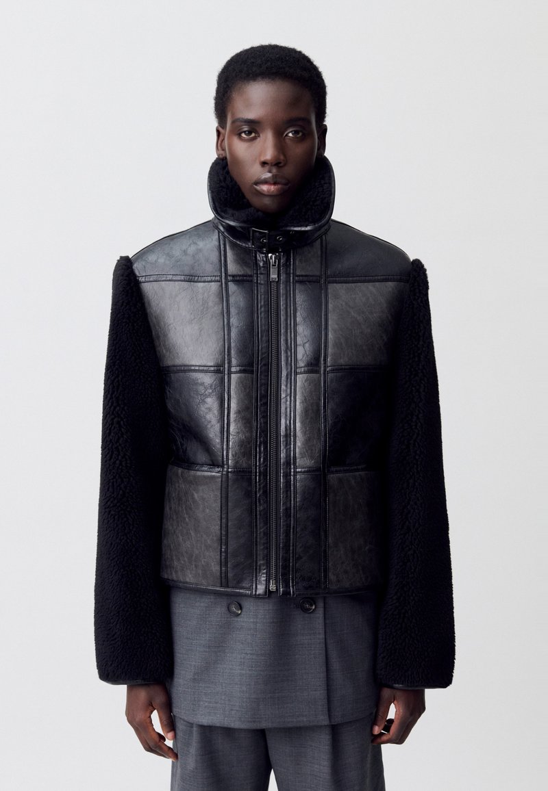 Chaqueta de cuero negro con un diseño acolchado, que cuenta con mangas de tela negra rizada y un cuello alto. Cierre de cremallera en la parte delantera.