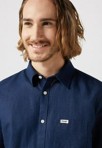 Camicia blu navy con bottoni, maniche lunghe, colletto e bottoni bianchi. Piccola etichetta bianca con "Wrangler" sulla tasca sinistra. Texture liscia.