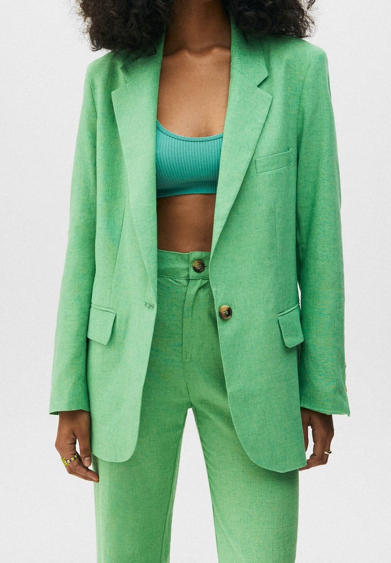 Groene linnen broekpak met een op maat gemaakte blazer met twee knopen, voorzakken en bijpassende taillepants. Geribbelde turquoise crop top eronder.
