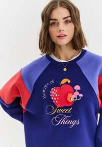 Niebieska i czerwona bluza z grafiką czerwonego jabłka z grzybami oraz napisem "Uważaj na słodkie rzeczy." Wykonana z mieszanki bawełny.