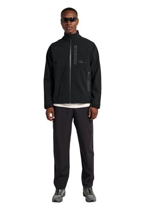 Schwarze Softshelljacke mit Reißverschlusstaschen, hohem Kragen und Logo-Detail, kombiniert mit schwarzen Hosen und grauen Turnschuhen. Sonnenbrille getragen.