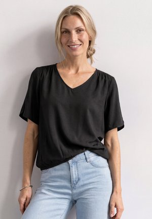 Smilende kvinde med blondt hår iført en løs sort V-hals bluse og lyseblå jeans står foran en ensartet lys baggrund.