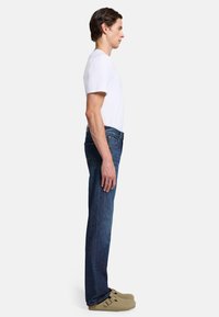 Slim-fit blå denimjeans med bakfickor, bärs med en vit t-shirt med rund halsringning och beigefärgade slip-on skor, sedd från sidan.