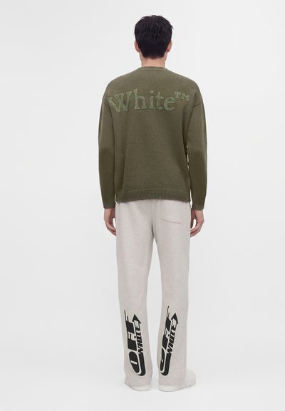 Homme debout, dos tourné, portant un pull vert olive avec le texte "White" et un pantalon clair avec des logos noirs "Off-White" à l'arrière des jambes.