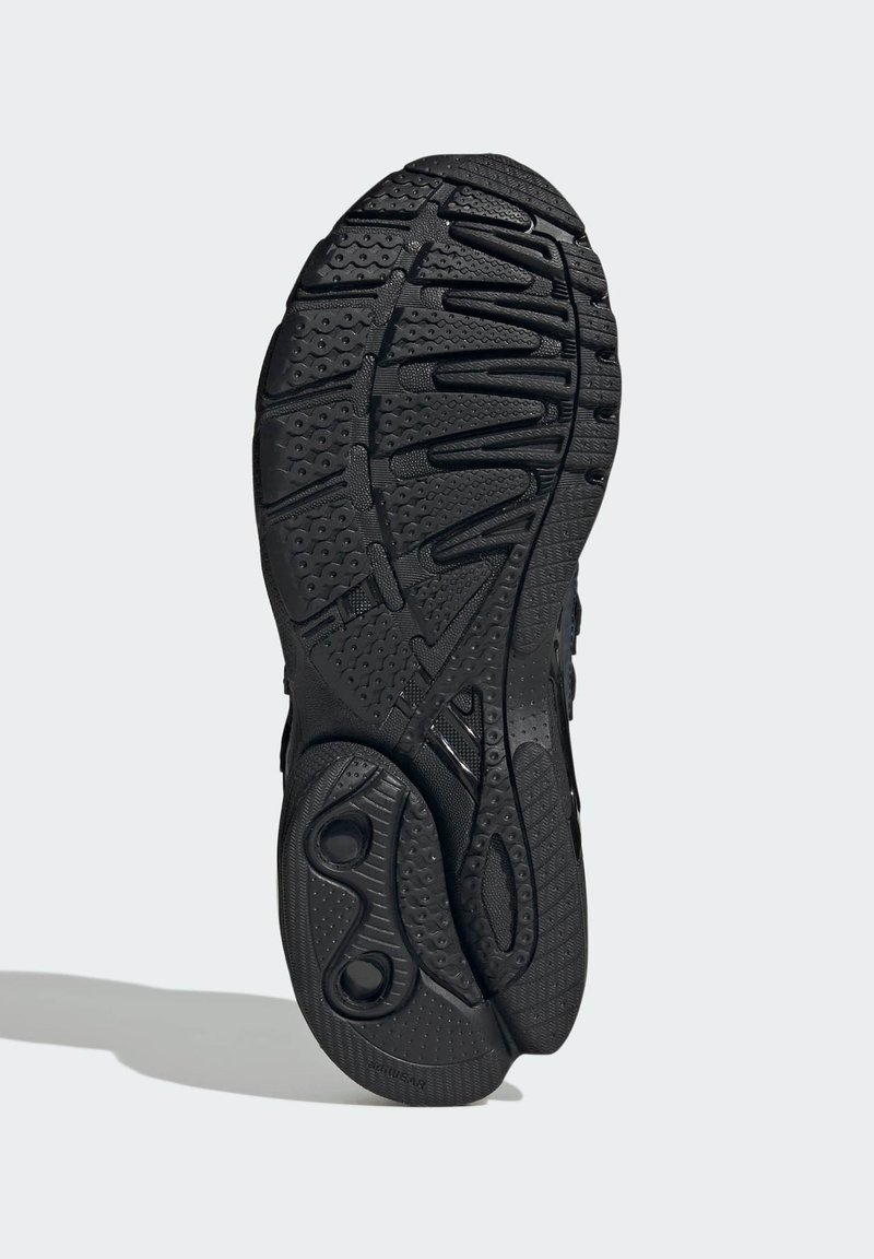 Sort sneakersål med tekstureret, opdelt slidmønster designet til greb og fleksibilitet, vist mod en ensartet lys baggrund.