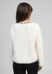 Pull en tricot blanc avec un motif texturé, ornée d'une bordure festonnée noire le long de l'encolure et de l'ourlet, porté avec un pantalon noir.
