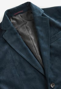 Blazer en daim bleu marine avec un revers en pointe, fermeture à un bouton et doublure grise à motifs. Il présente des détails cousus et une poche poitrine.